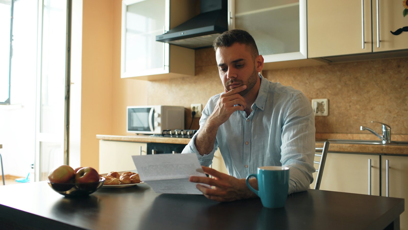 A man at a kitchen table reading a letter with a serious look