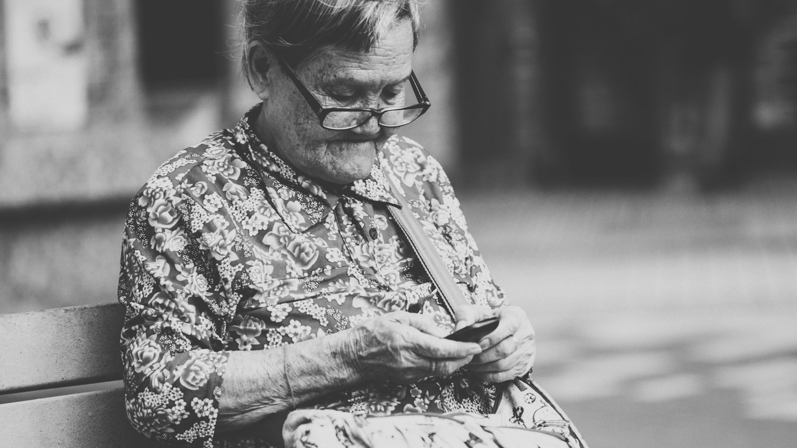 A black and white photo of an older woman looking at her phone on a bench
