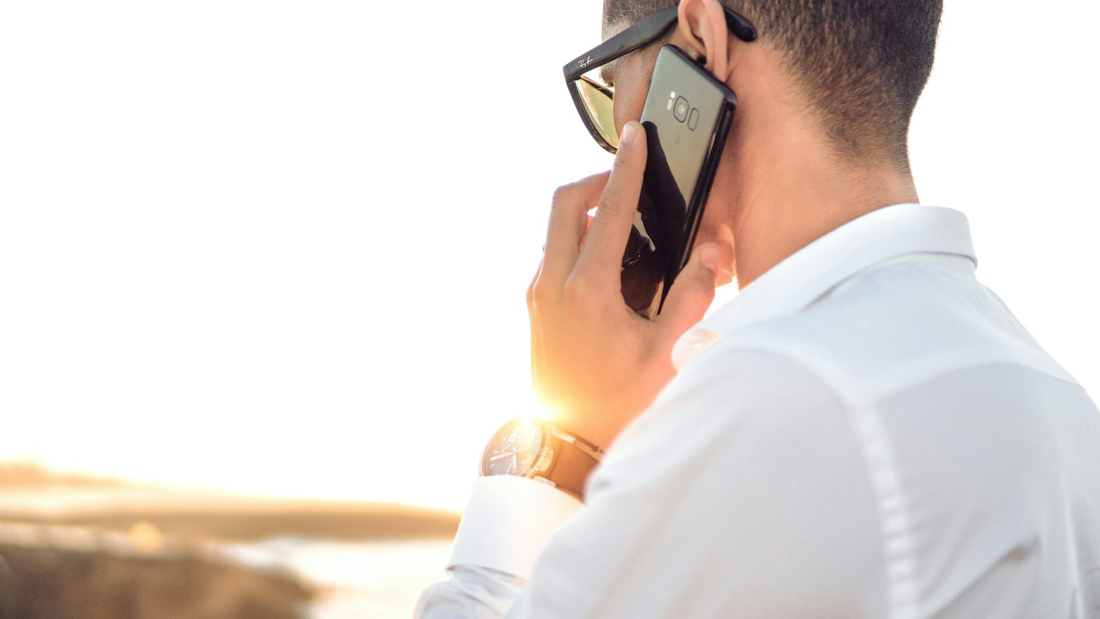 A man in a white shirt on a phone call by the water at sunset