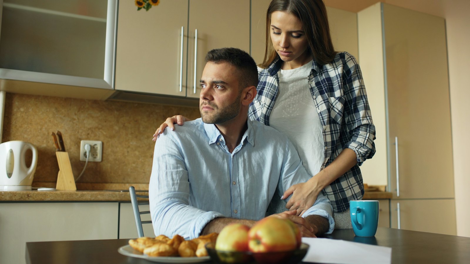 A woman comforting a man at a kitchen table
