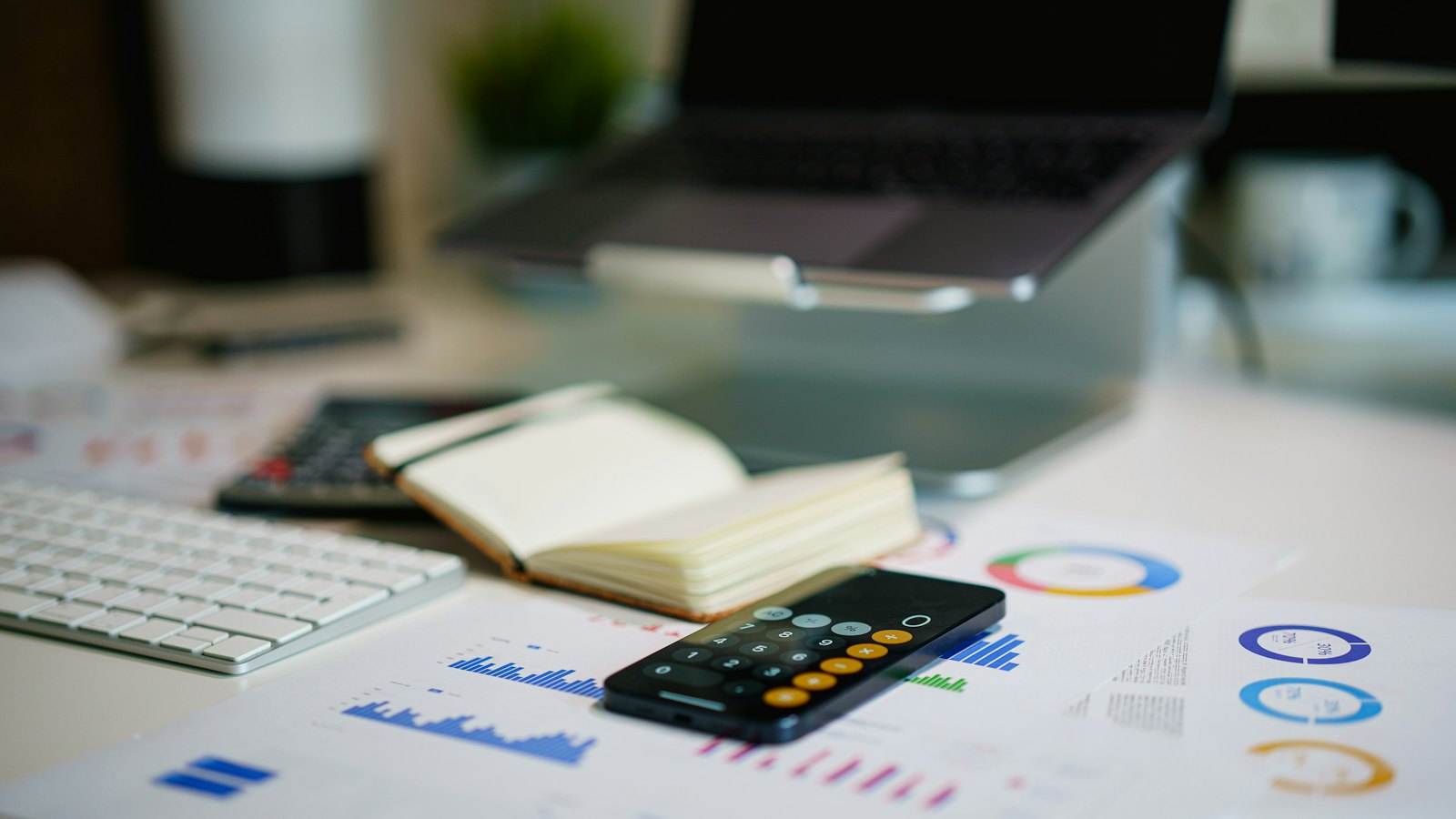 A finance desk with a calculator, a notebook and printed charts beside a laptop