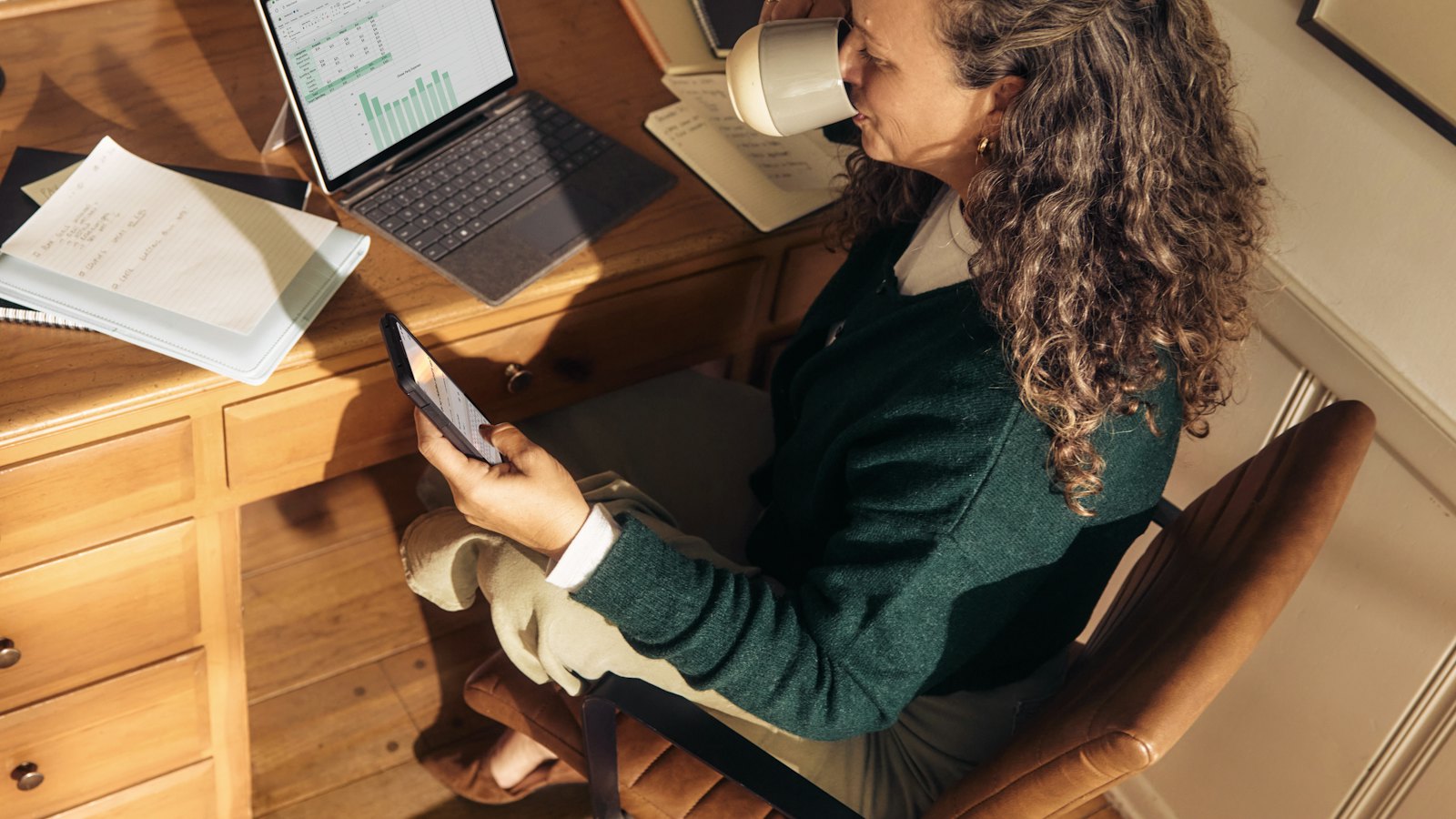 A woman at a home desk checking her phone beside a laptop