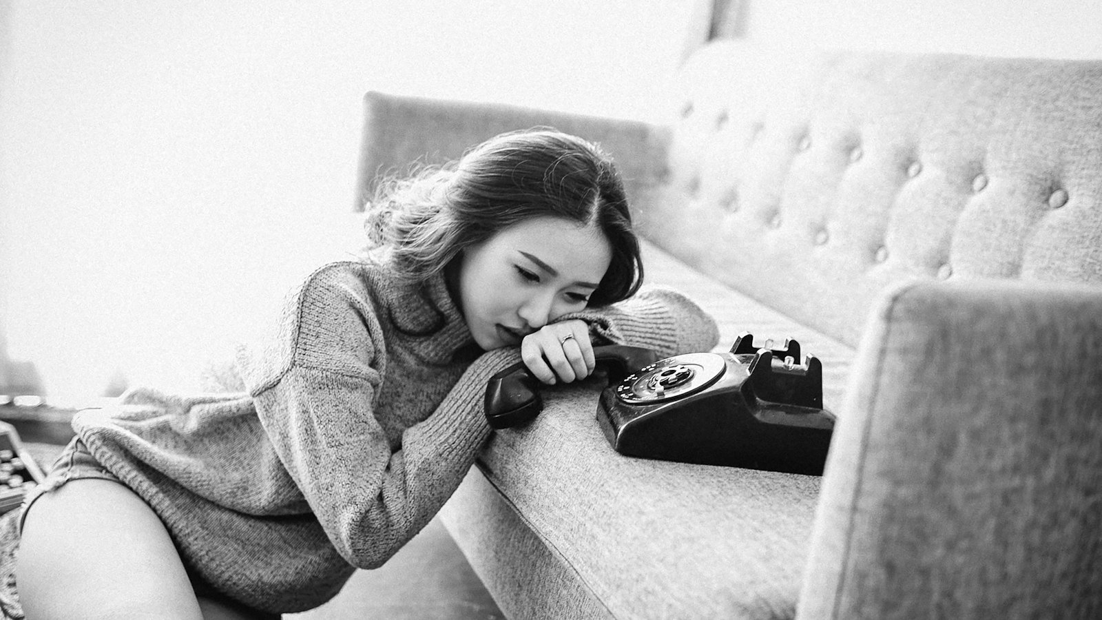 A black and white photo of a woman resting her head on her arm beside an old telephone