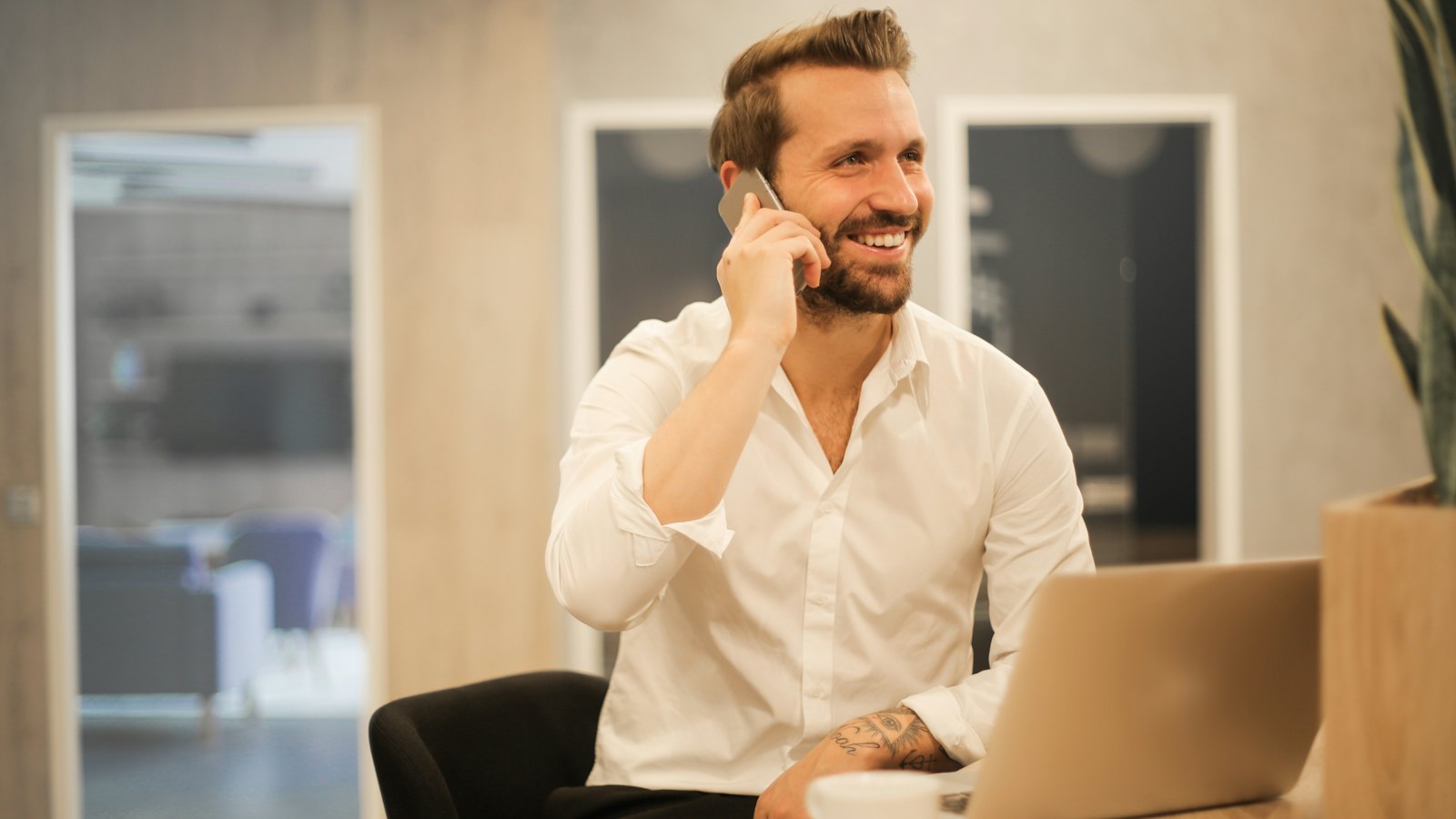 A smiling man in a white shirt on a phone call at his desk