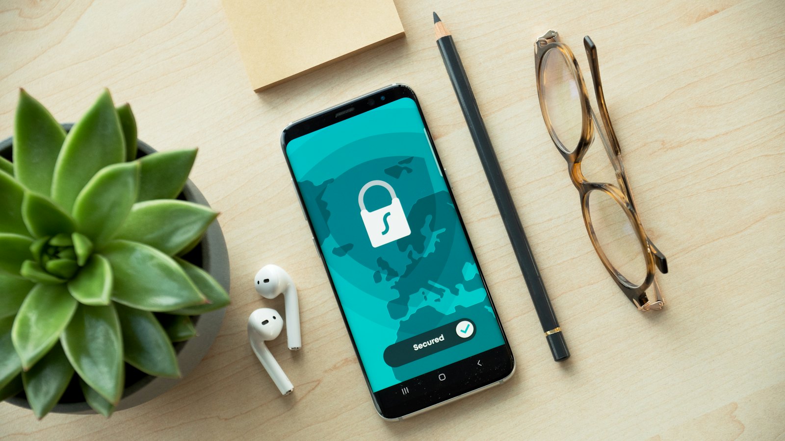 A phone on a desk showing a padlock and the word Secured, beside a plant, earphones and glasses