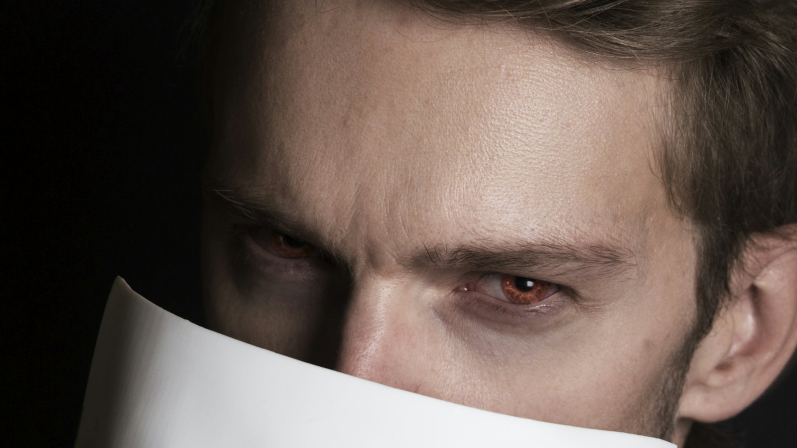 A man half hiding his face behind a sheet of paper, only his eyes visible