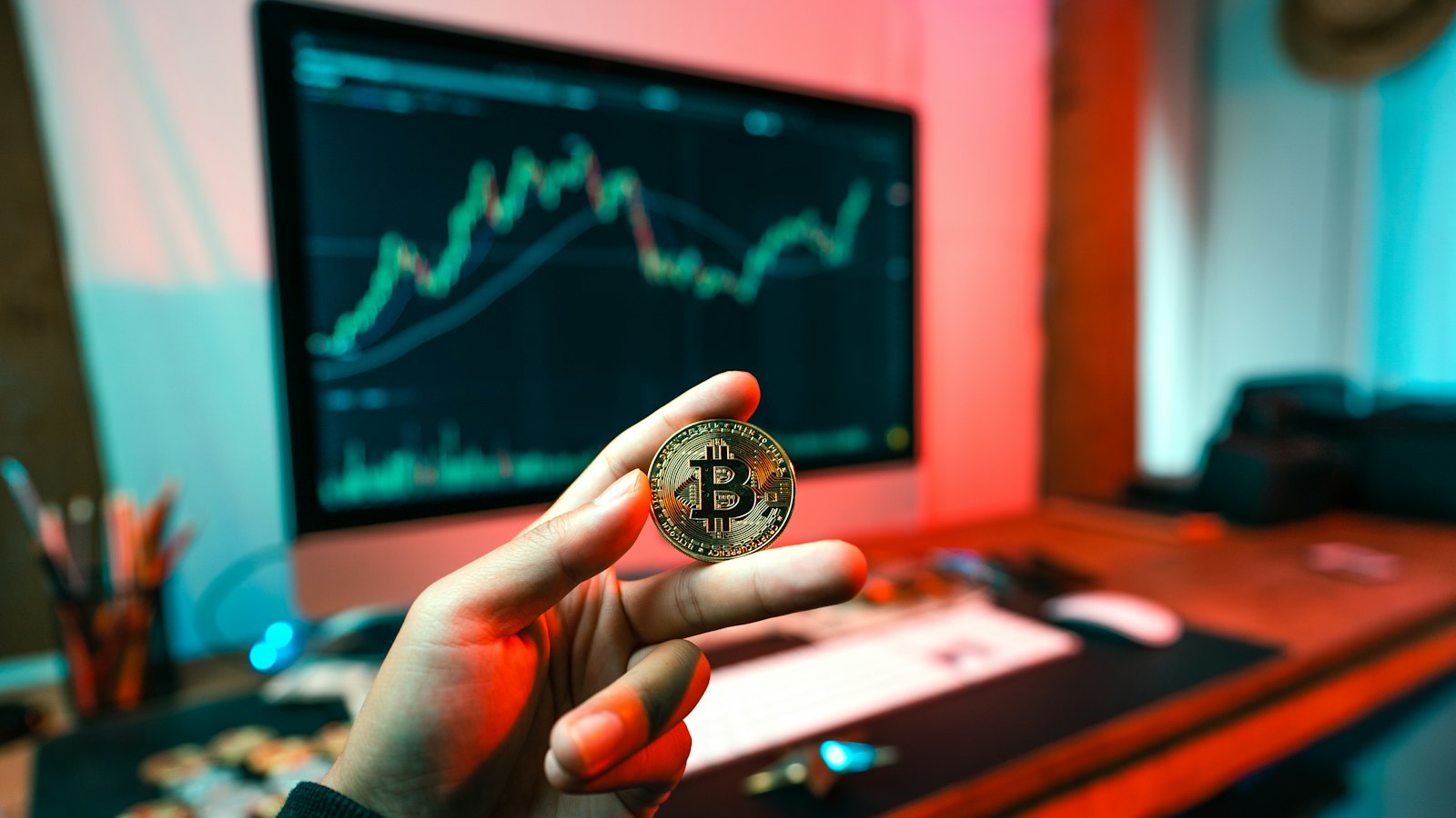 A hand holding a coin in front of a trading screen