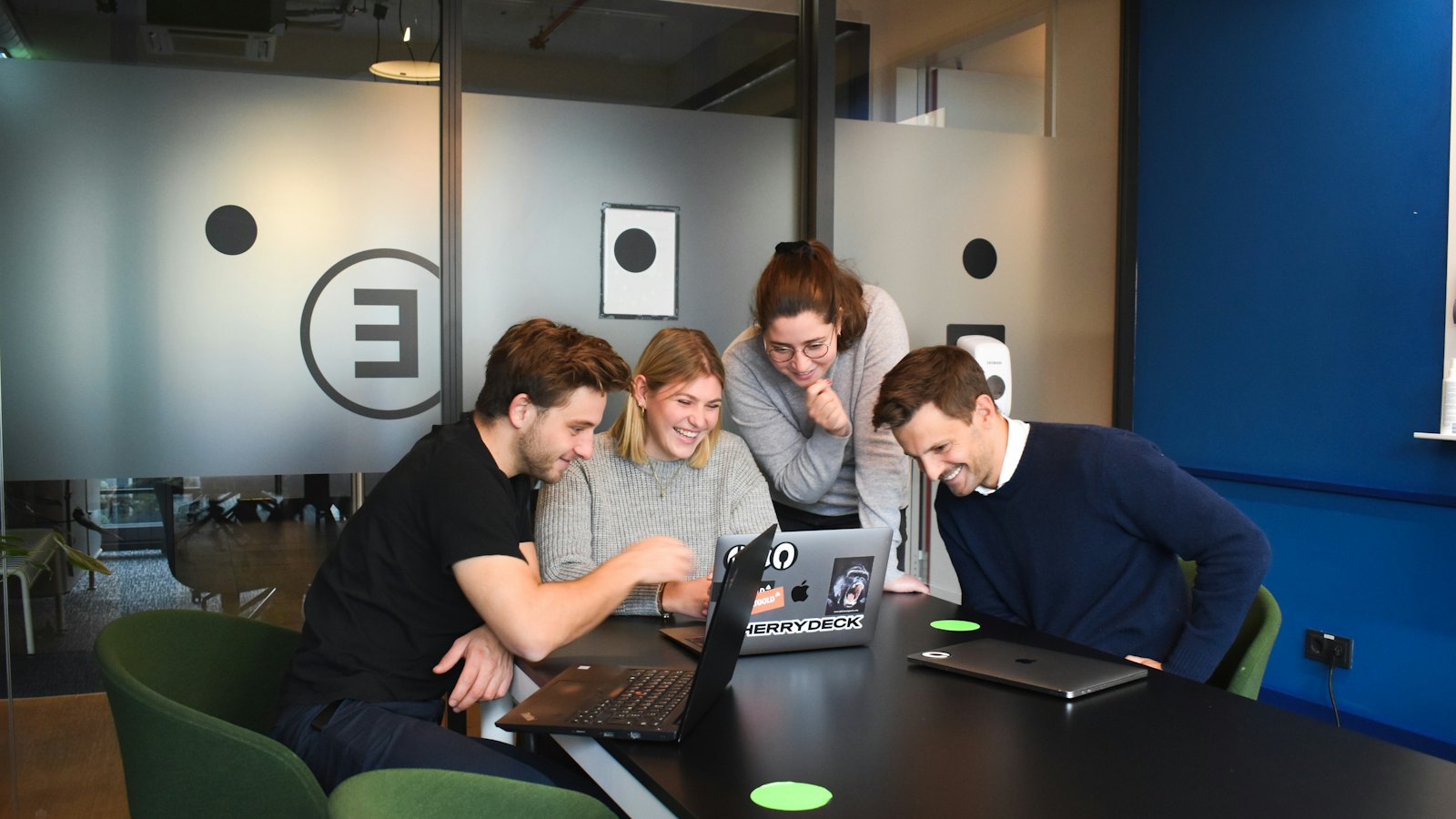 Four colleagues gathered around a laptop in a meeting room, reacting to what is on screen