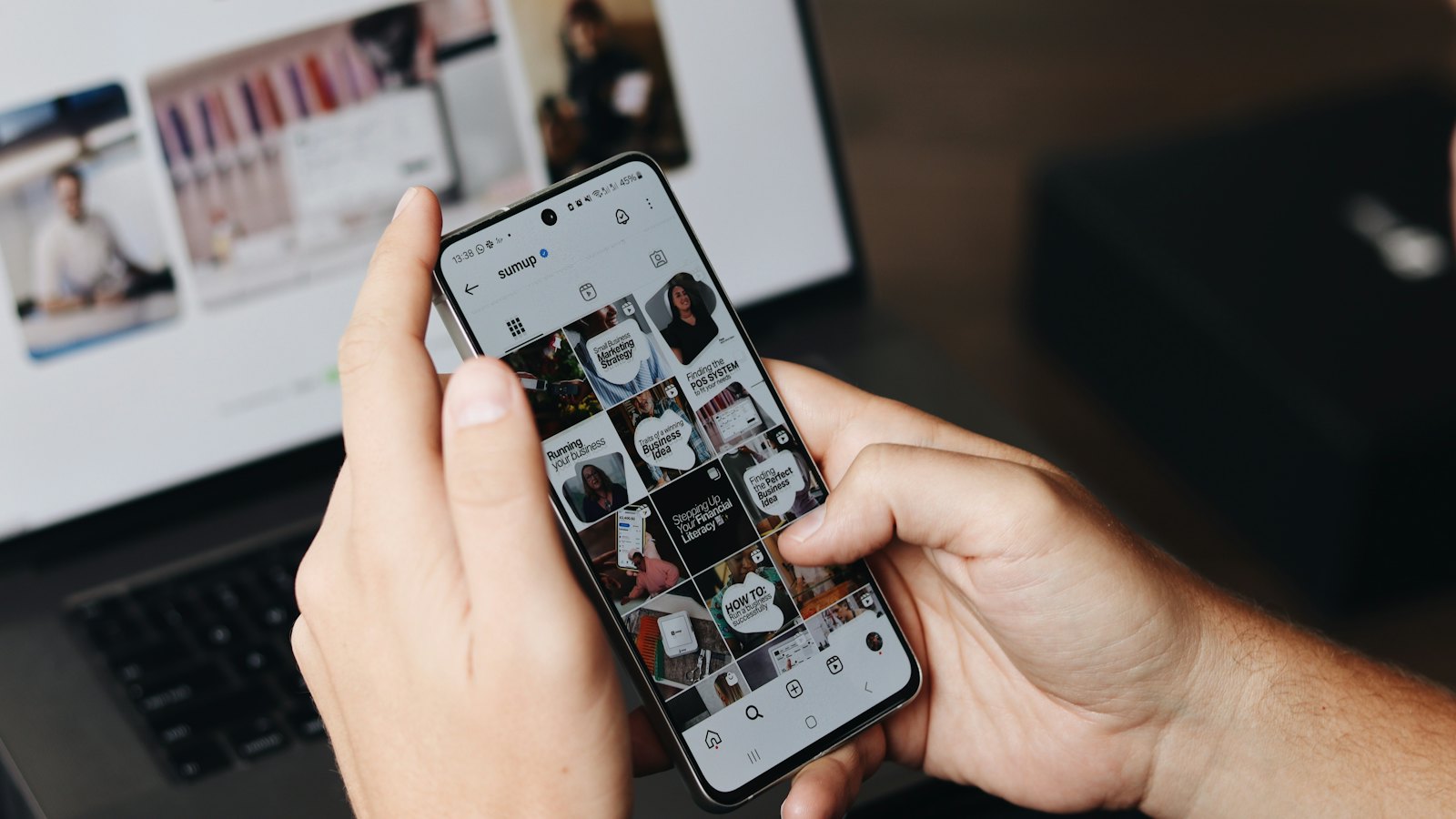 A person browsing a social media profile on a phone beside a laptop