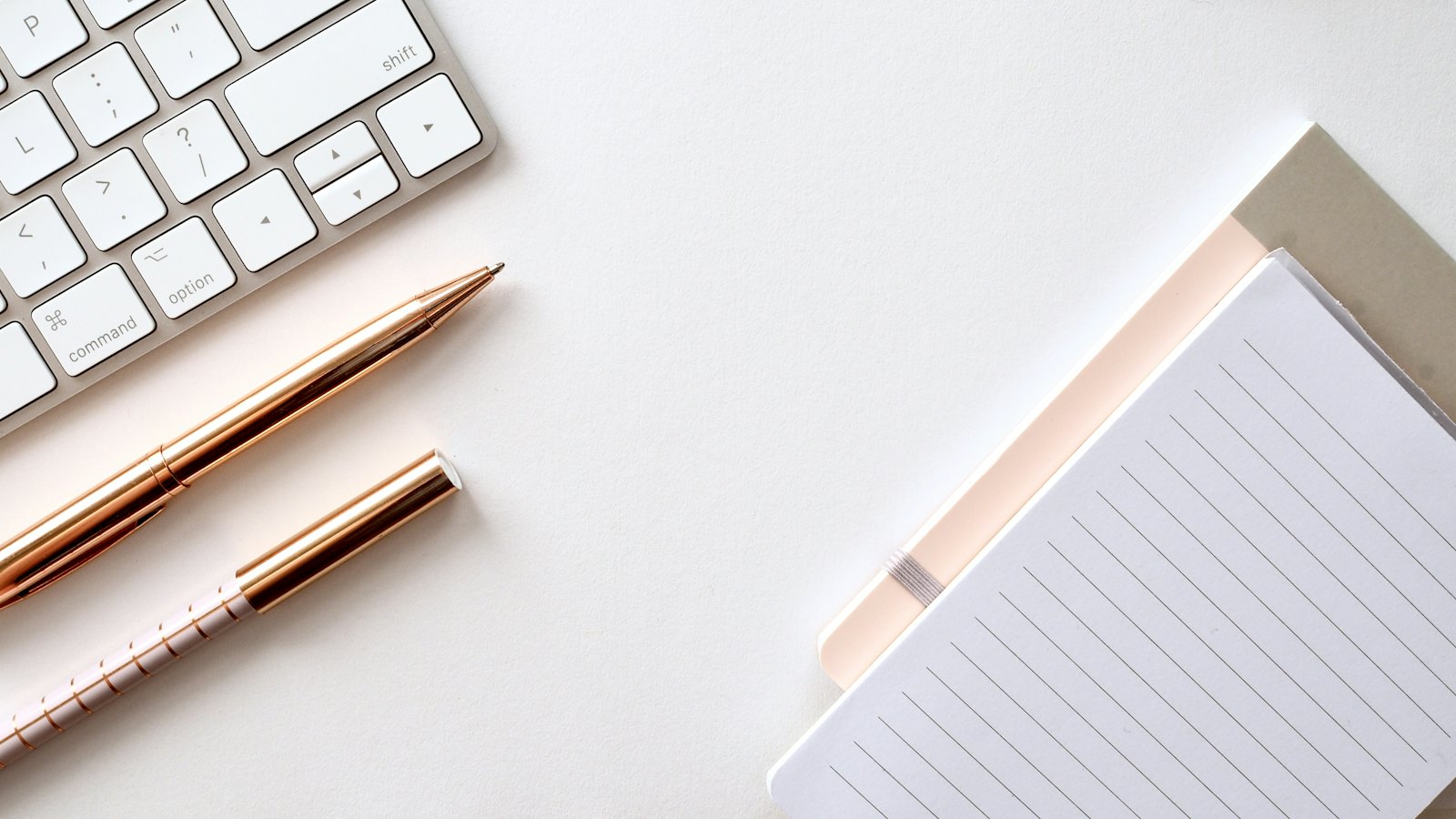A keyboard, pens and a notepad on a white desk