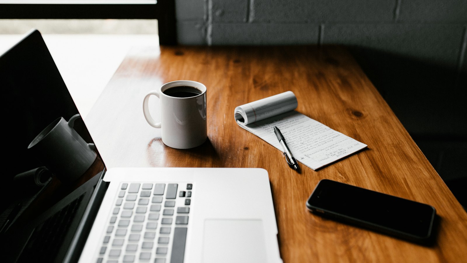 A laptop, a phone, a coffee cup and a notepad on a wooden desk