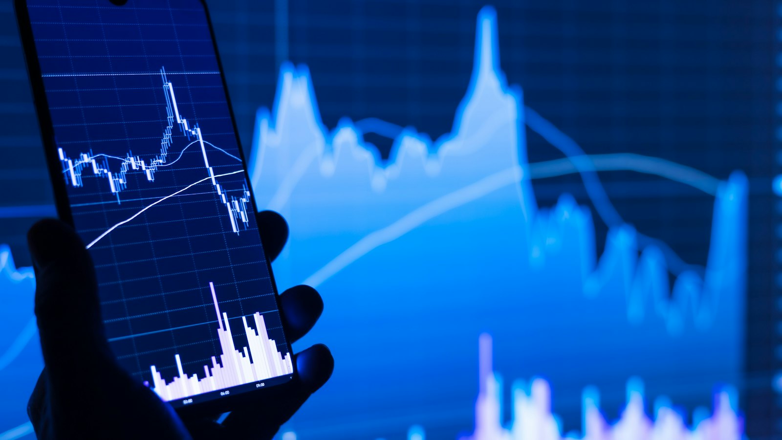 A hand holding a phone showing a trading chart in front of a larger chart