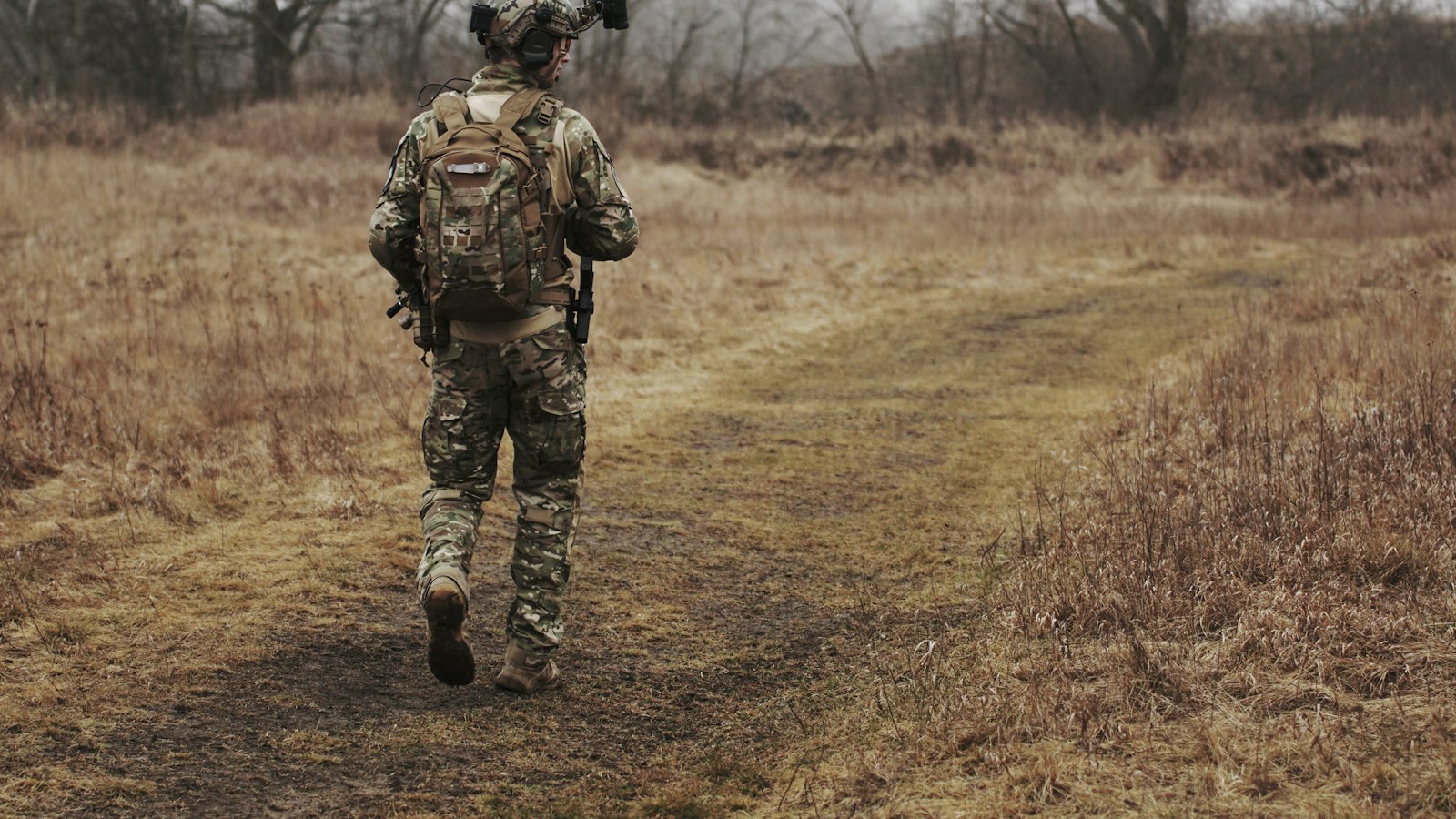 A soldier in camouflage with a backpack walking along a dirt path