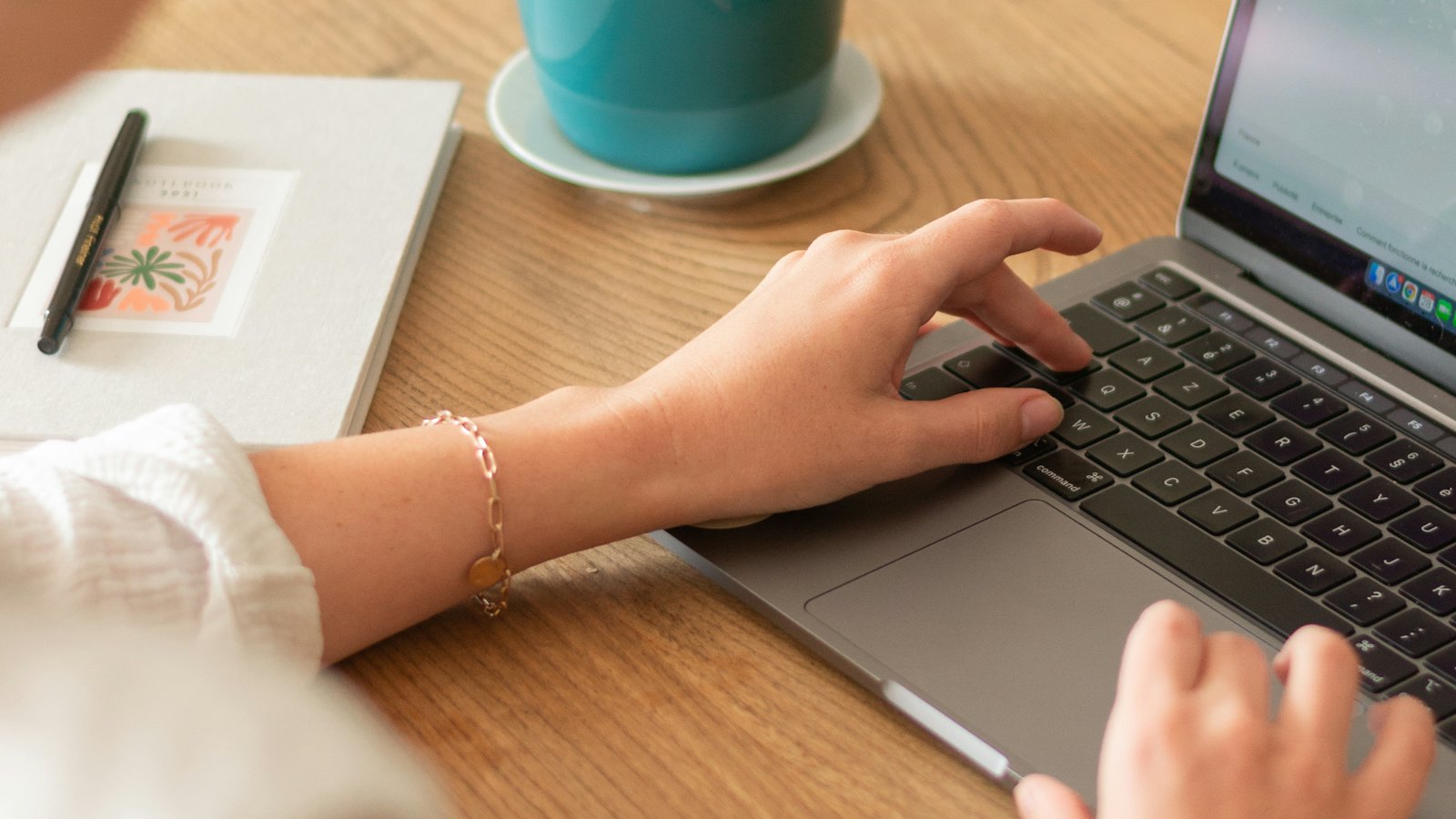 A hand typing on a laptop at a wooden table beside a cup