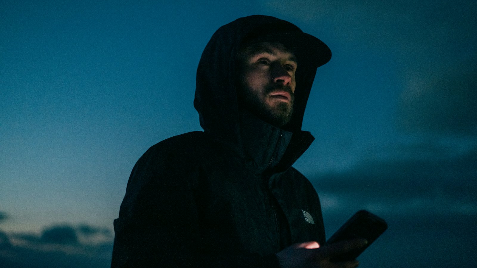 A person in a hood holding a phone at dusk