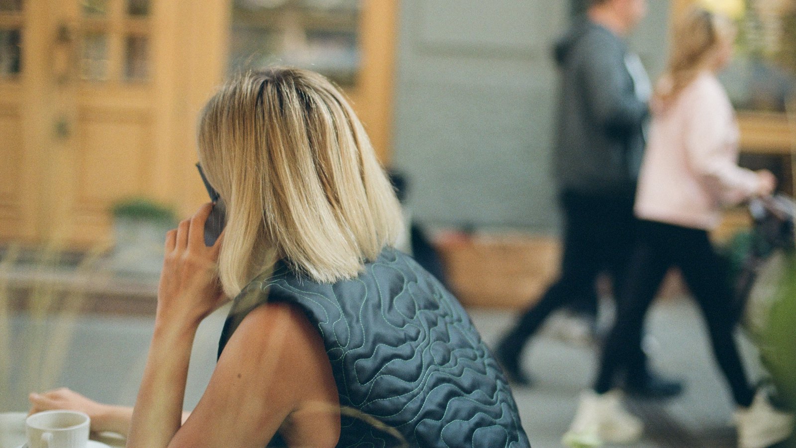 A woman on a phone call at a cafe table, with people walking past behind her