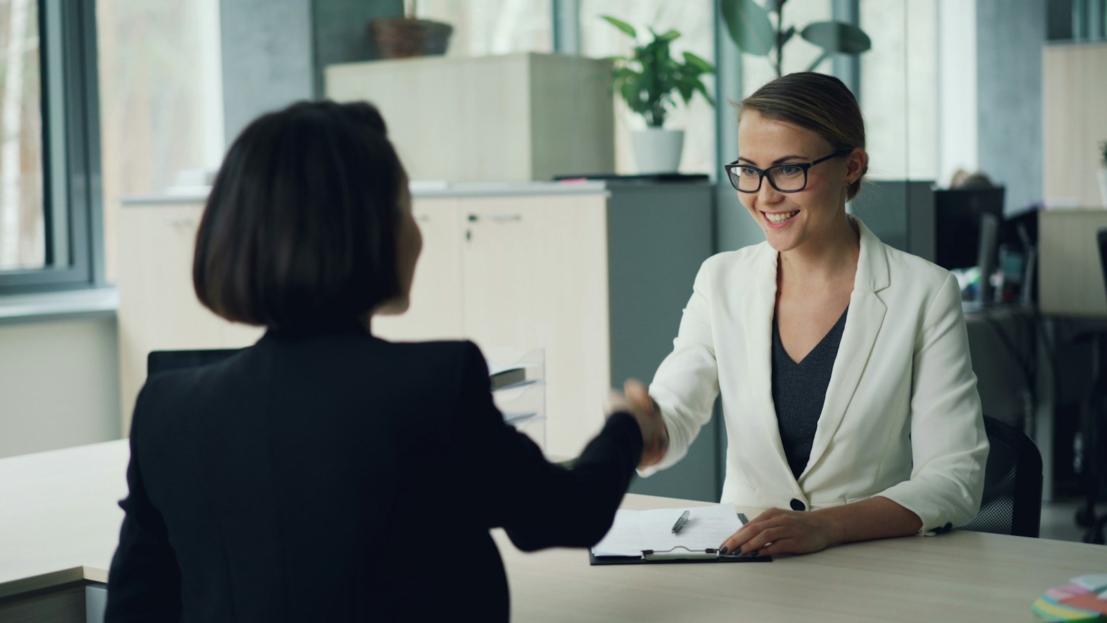 Two women shaking hands across a desk in an office