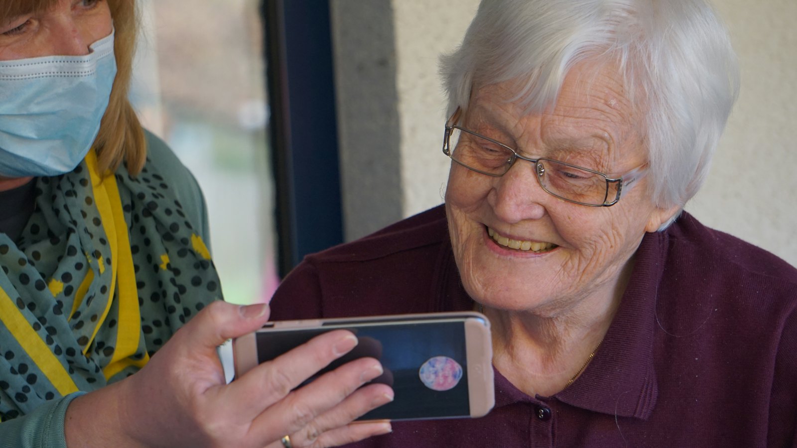 An older woman smiling at a message on a phone held by a friend