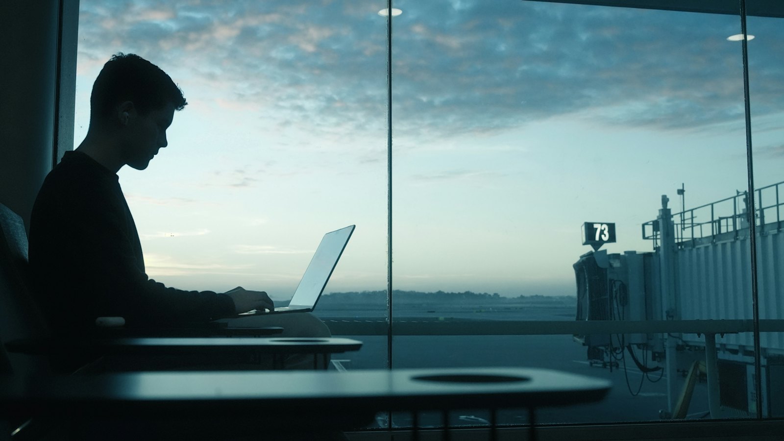 A man working on a laptop by a large airport window at dawn